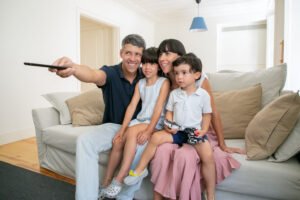 Happy parent couple with two kids watching TV, sitting on couch in living room and using remote control. Medium shot. Leisure time with family or television concept